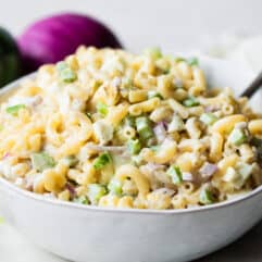 White bowl filled with macaroni salad with a lemon wedge on the side, green bell pepper and red onion in the background.