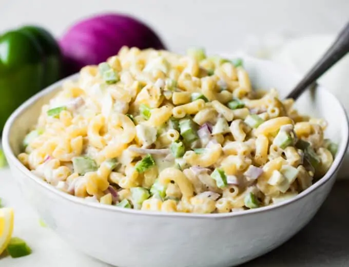 White bowl filled with macaroni salad with a lemon wedge on the side, green bell pepper and red onion in the background.