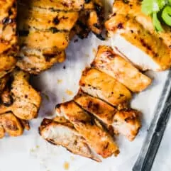 Sliced Mexican grilled chicken on a cutting board with parchment paper, cilantro and a knife.