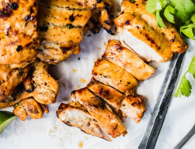 Sliced Mexican grilled chicken on a cutting board with parchment paper, cilantro and a knife.