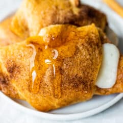 Baked sopapilla cheesecake rolls on a white plate with a drizzle of honey dripping down, a honey stirrer stick to the side.
