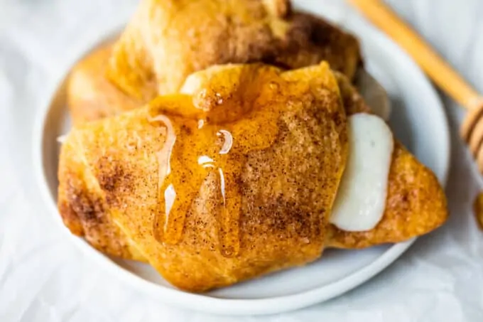 Baked sopapilla cheesecake rolls on a white plate with a drizzle of honey dripping down, a honey stirrer stick to the side.