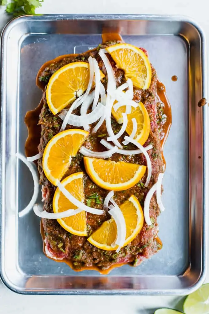 Overhead view of flank steak smothered in carne asada marinade, topped with orange slices and sliced onion.