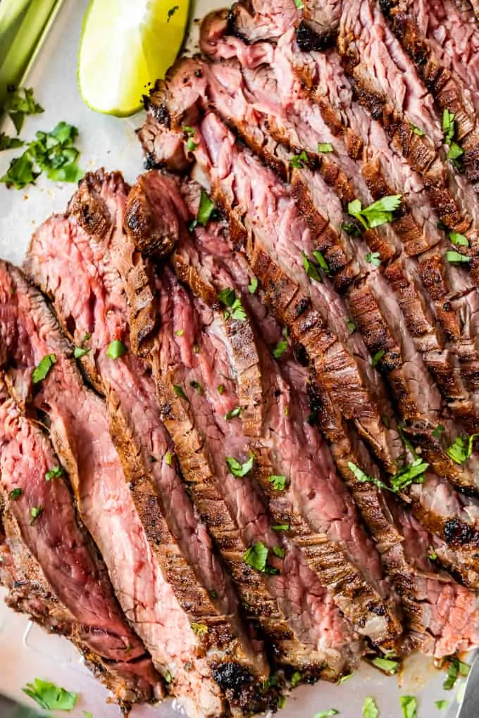 Overhead view of flank steak that has been marinated in carne asada marinade, and grilled. Sliced and laid out on a baking sheet. Topped with bits of green diced cilantro and a lime wedge on the side.