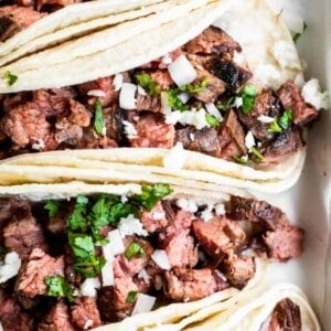 Overhead view of layered white corn tortillas that are filled with chopped grilled carne asada, topped with onion and cilantro, and crumbled queso fresco.