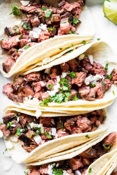 Carne Asada tacos lined up and topped with diced onion, cilantro and crumbled queso fresco. Lime wedges on the side.