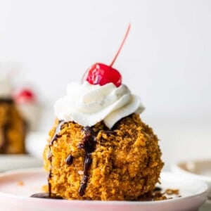 Easy to make mexican fried ice cream on a pink plate topped with drizzles of chocolate fudge, whipped cream and a cherry on top.