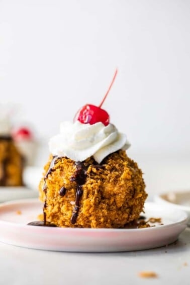 Easy to make mexican fried ice cream on a pink plate topped with drizzles of chocolate fudge, whipped cream and a cherry on top.