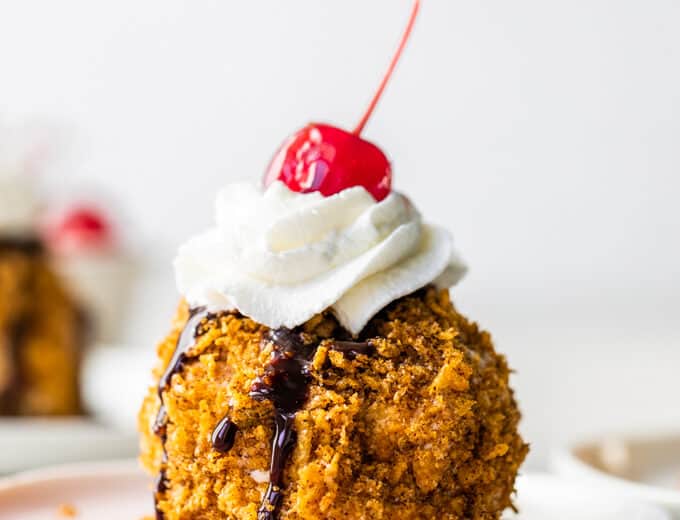Easy to make mexican fried ice cream on a pink plate topped with drizzles of chocolate fudge, whipped cream and a cherry on top.
