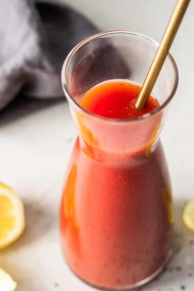 A carafe filled with homemade Clamato juice with a golden spoon to stir and lemon wedges on the side.