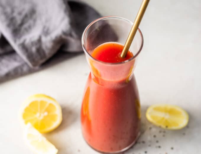 A carafe filled with homemade Clamato juice with a golden spoon to stir and lemon wedges on the side.