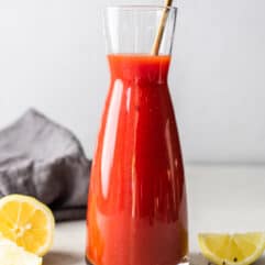 Carafe filled with homemade Clamato juice, red liquid, showing black pepper and seasonings. Lemon wedges lay on the counter around it, along with coarse black pepper.