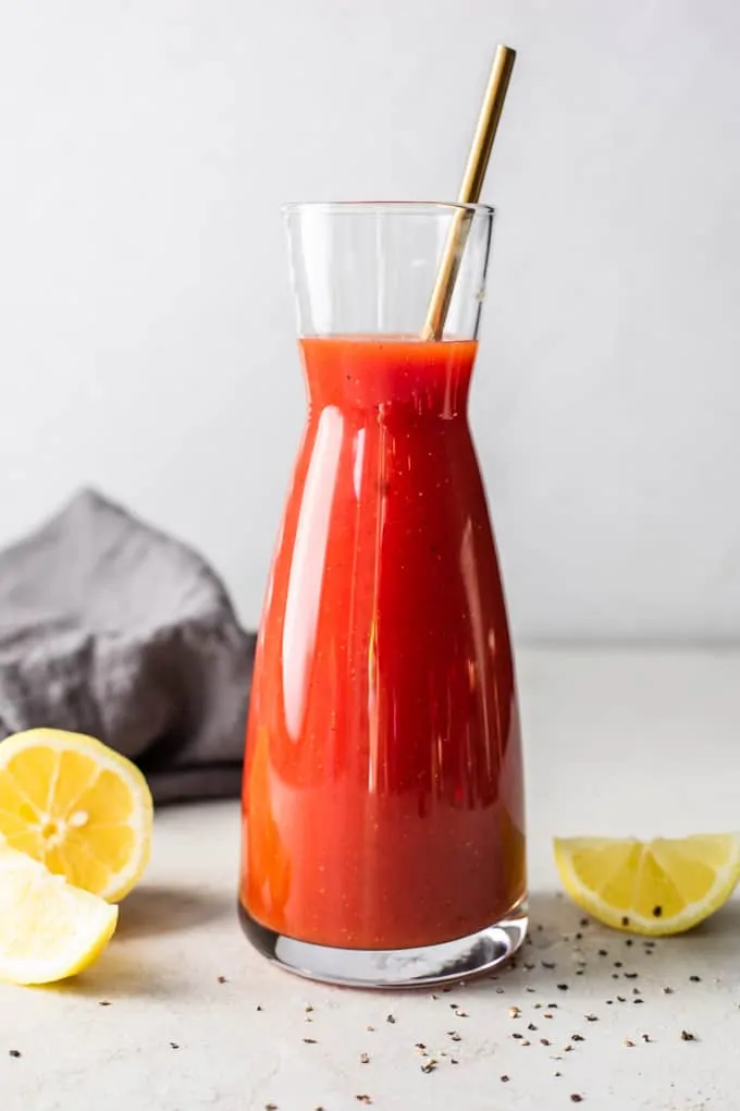 Carafe filled with homemade Clamato juice, red liquid, showing black pepper and seasonings. Lemon wedges lay on the counter around it, along with coarse black pepper. 