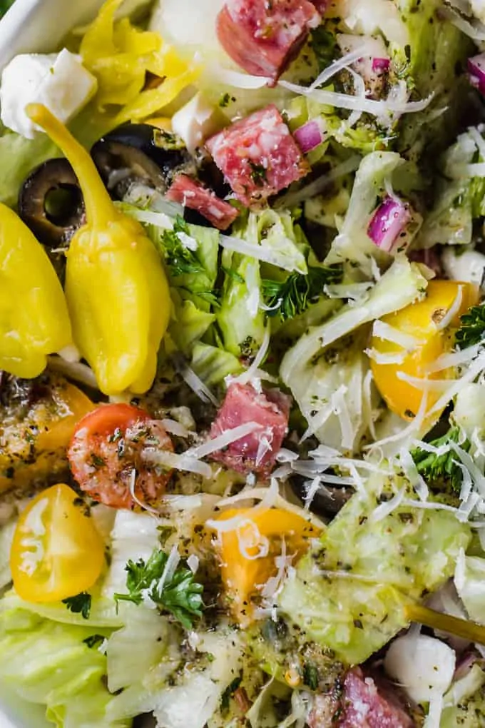 Up close view of Italian chop salad, lettuce with diced salami, tomatoes, chick peas, tomatoes, red onion, and more all drizzled with Italian dressing, topped with whole pepperoncinis.