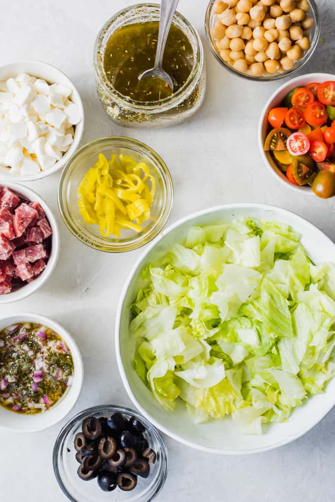 Ingredients laid out for an Italian Chop salad. A large bowl of chopped lettuce, salami, olives, tomatoes, chick peas, mozzarella, pepperoncinis, red onion, and a jar of homemade Italian dressing.