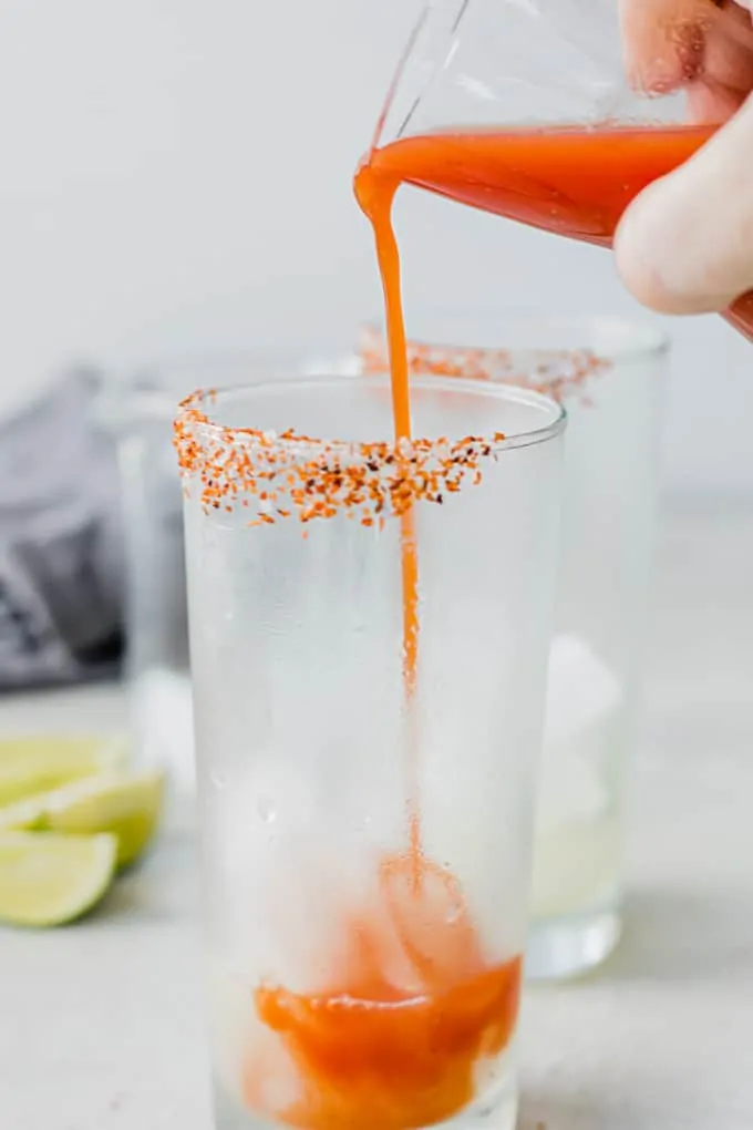 Clamato juice being poured into a frosty glass with Tajin on the rim.