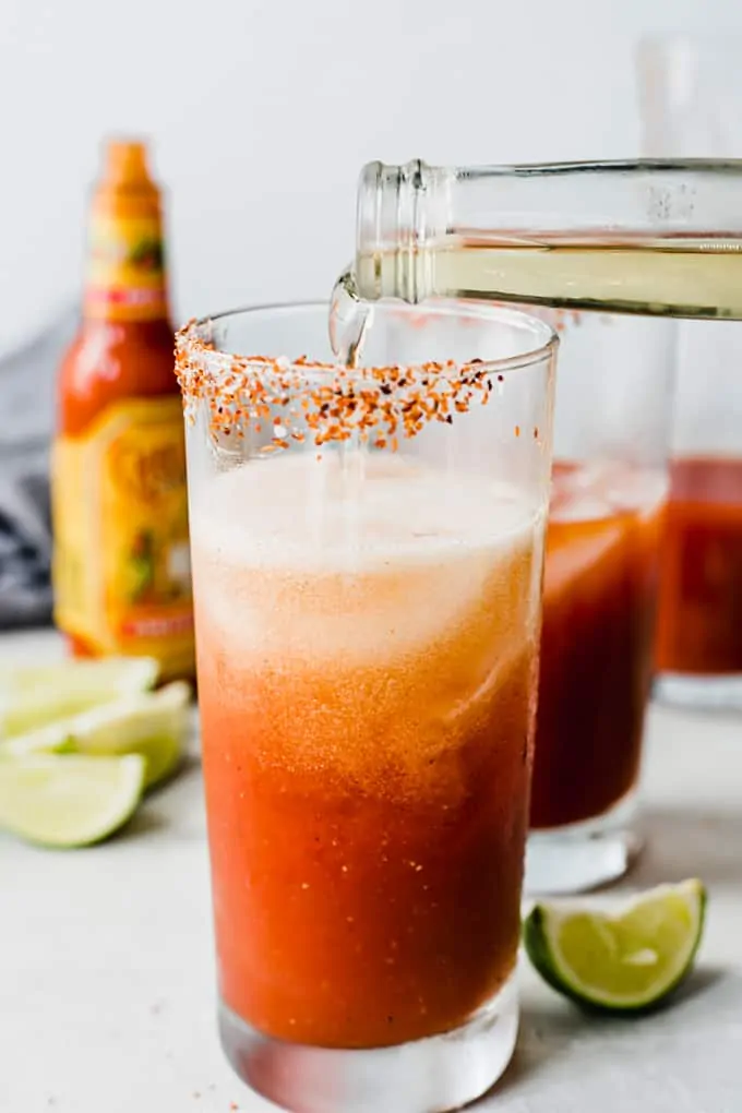Frosty mug with Tajin on the rim filled with ice and tomato juice, with a beer being poured in. 