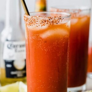 Glass filled with red Michelada drink, showing ice cubes, Tajin on the rim of the cup, lime wedges on the counter, and a corona bottle behind the glasses.