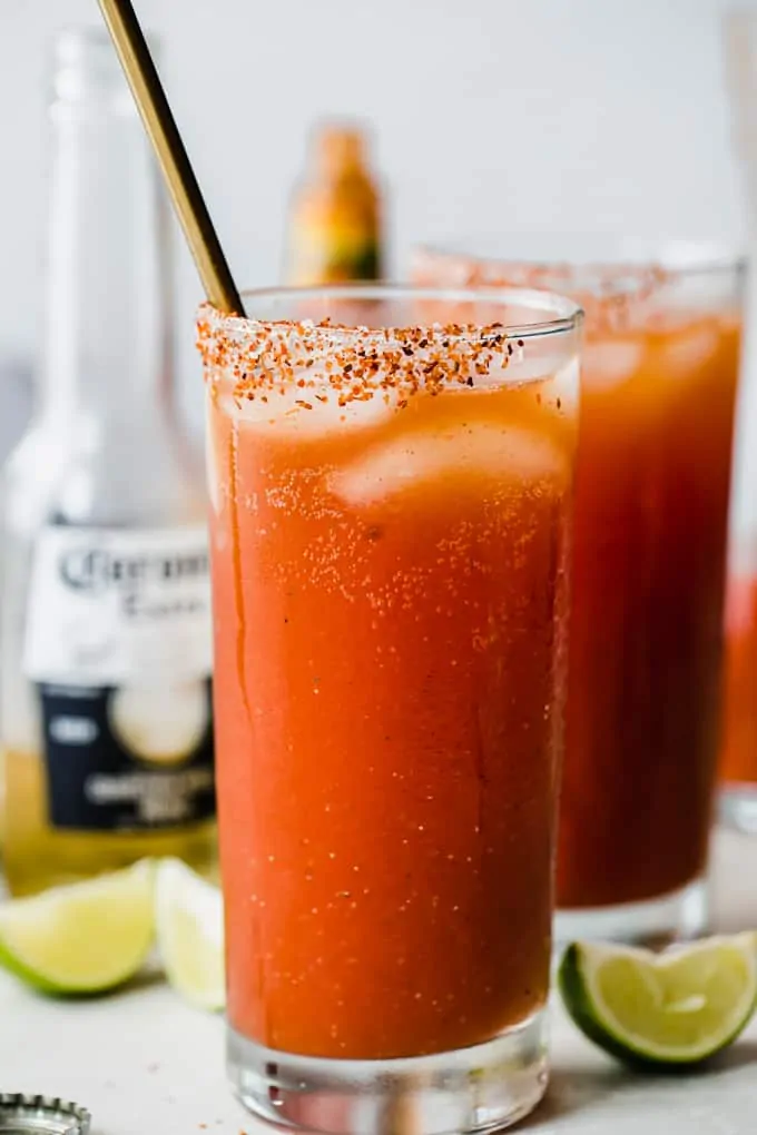 Glass filled with red Michelada drink, showing ice cubes, Tajin on the rim of the cup, lime wedges on the counter, and a corona bottle behind the glasses.