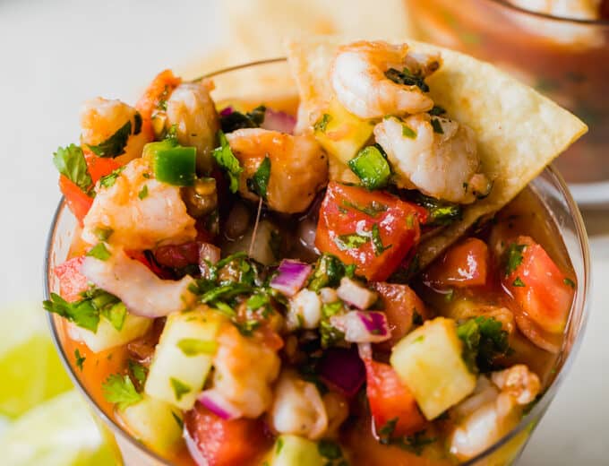 Cup filled with shrimp ceviche, showing red juice, chunks of shrimp, cucumber, tomato, jalapeno, red onion and cilantro. Lime wedges on the side, tortilla chips, and cilantro.