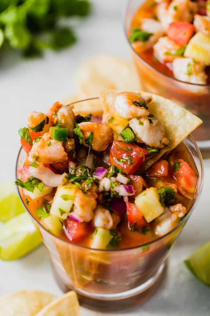 Cup filled with shrimp ceviche, showing red juice, chunks of shrimp, cucumber, tomato, jalapeno, red onion and cilantro. Lime wedges on the side, tortilla chips, and cilantro.