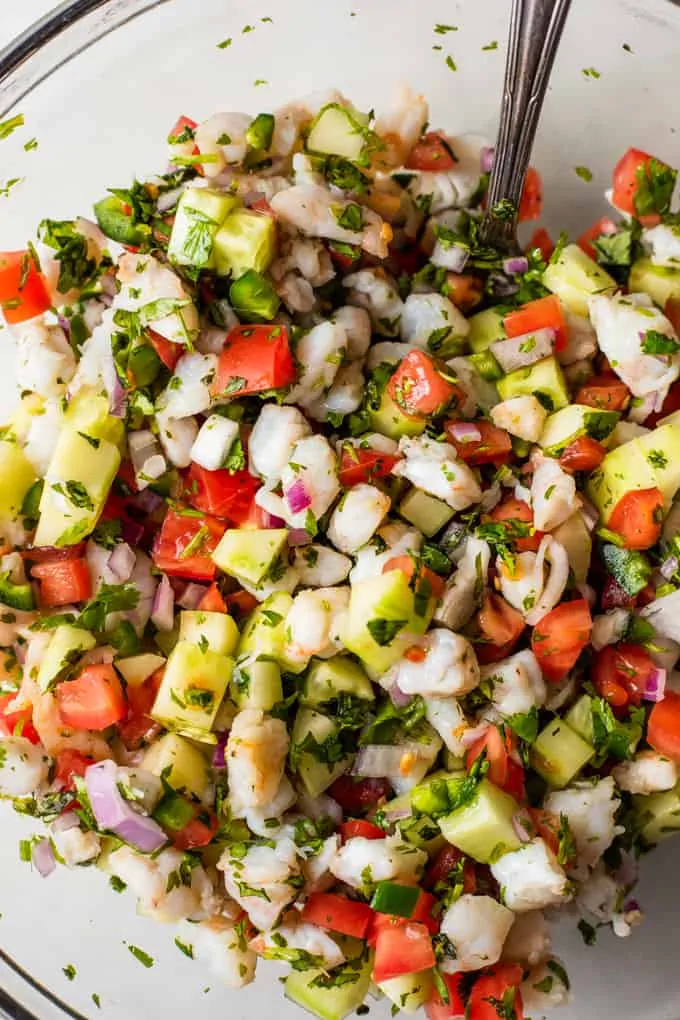 Glass bowl filled with chopped shrimp, cucumber, tomato, jalapeno, red onion and cilantro.