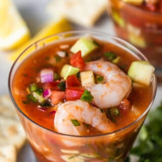Small cocktail glass filled with Mexican shrimp cocktail, showing shrimp, tomatoes, avocado, red onion, and cucumber swimming in a red spiced juice.