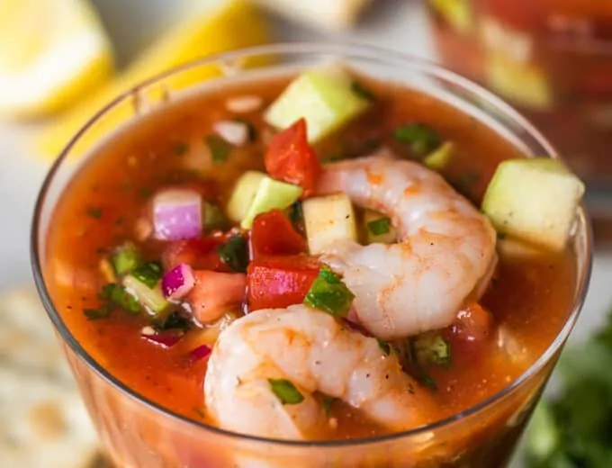 Small cocktail glass filled with Mexican shrimp cocktail, showing shrimp, tomatoes, avocado, red onion, and cucumber swimming in a red spiced juice.
