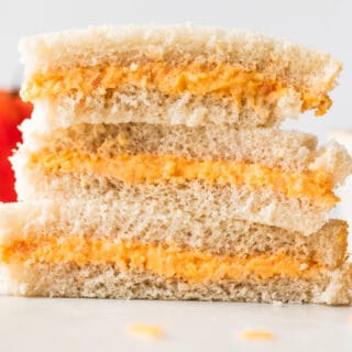 Stack of tomato twiddle sandwiches on white bread, with grated cheese on the counter around them, a tomato in the background and a ramekin filled with tomato twiddle.
