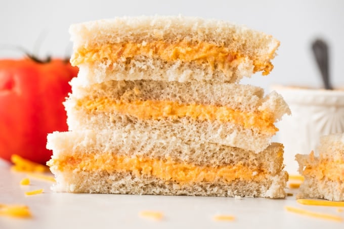 Stack of tomato twiddle sandwiches on white bread, with grated cheese on the counter around them, a tomato in the background and a ramekin filled with tomato twiddle.