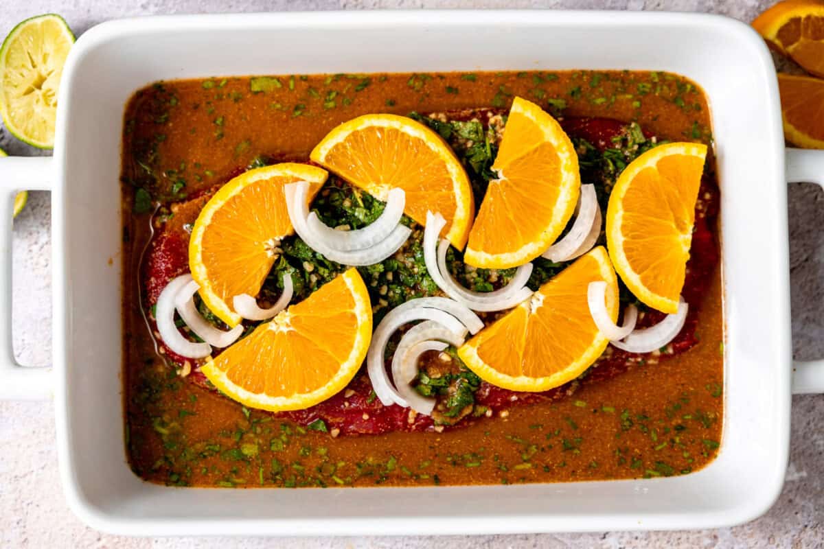 Flank steak being marinated in a carne asada marinade topped with orange slices and sliced onion. 