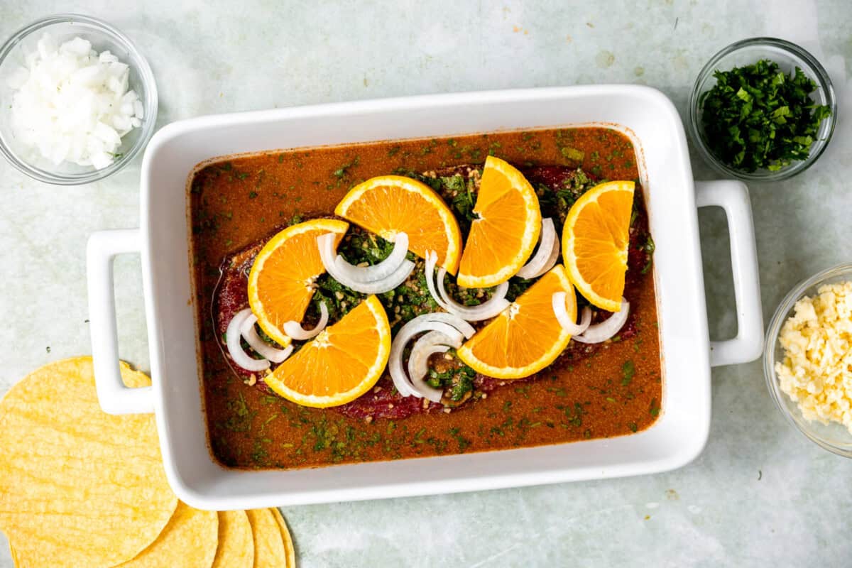 Carne Asada marinating in a baking dish topped with orange slices and sliced onion. 