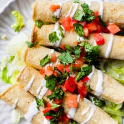 A plate filled with baked chicken taquitos, topped with drizzles of Crema, diced tomato, and fresh cilantro.