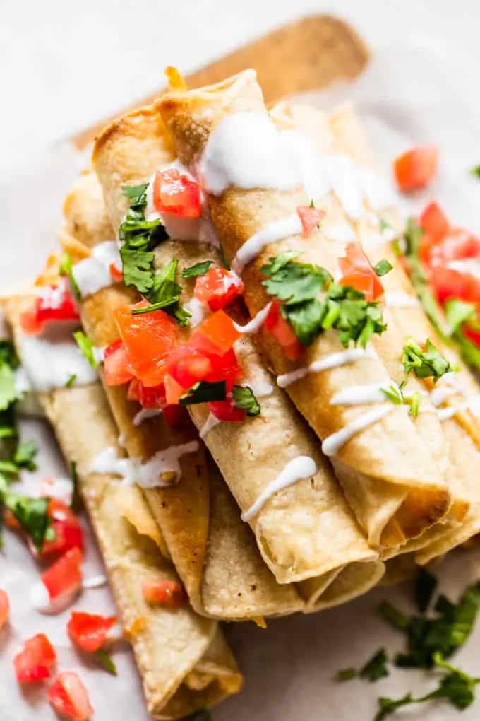 Overhead view of stacked taquitos drizzled with Crema, diced tomato and cilantro. 