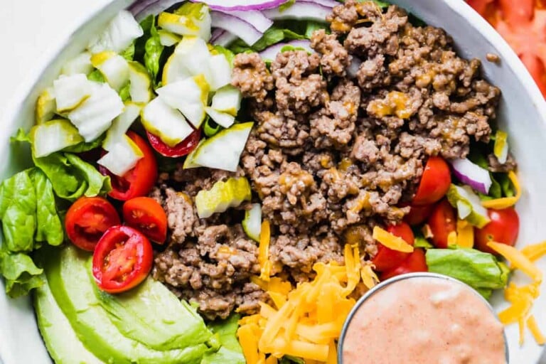 White bowl filled with chopped green lettuce, topped with cooked burger meat, cheese, avocado, tomato, pickle, red onion and a cup of burger sauce.