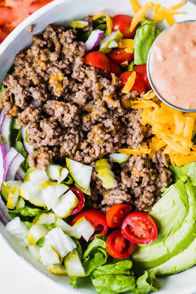 White bowl filled with chopped green lettuce, topped with cooked burger meat, cheese, avocado, tomato, pickle, red onion and a cup of burger sauce. 