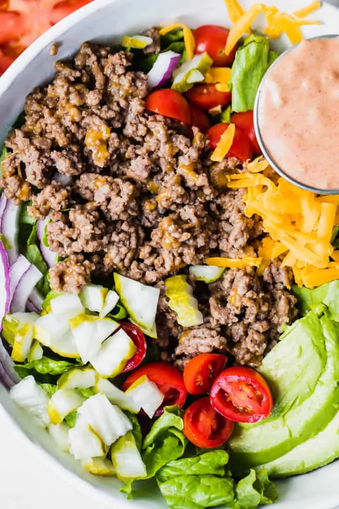 White bowl filled with chopped green lettuce, topped with cooked burger meat, cheese, avocado, tomato, pickle, red onion and a cup of burger sauce. 