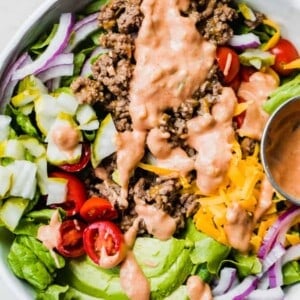 White bowl filled with chopped green lettuce, topped with cooked burger meat, cheese, avocado, tomato, pickle, red onion all drizzled with burger sauce.