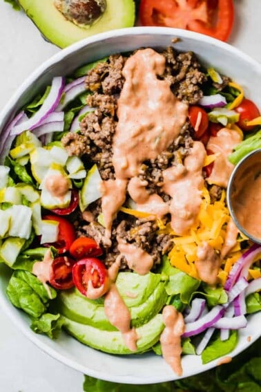 White bowl filled with chopped green lettuce, topped with cooked burger meat, cheese, avocado, tomato, pickle, red onion all drizzled with burger sauce.