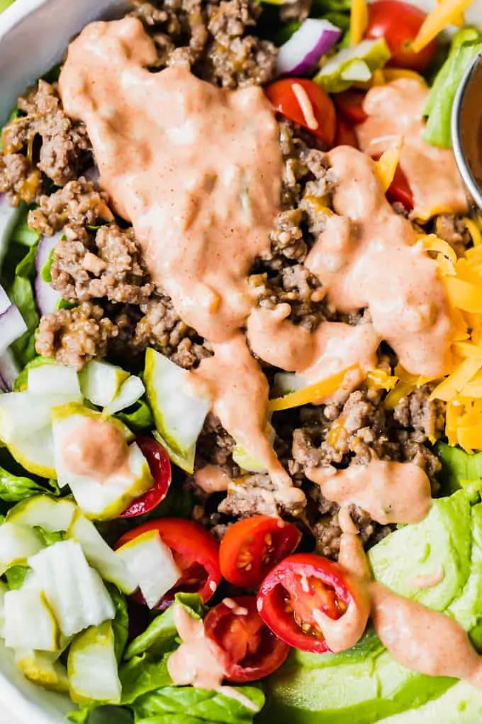 White bowl filled with chopped green lettuce, topped with cooked burger meat, cheese, avocado, tomato, pickle, red onion all drizzled with burger sauce.