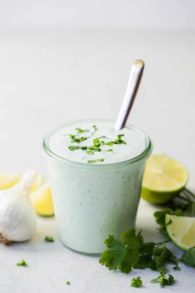 Jar filled with cilantro lime crema, surrounded by stems of cilantro, garlic, lime wedges.