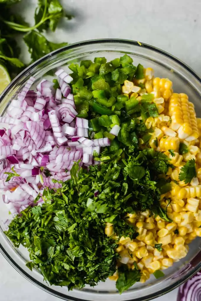 Glass bowl filled with ingredients to make homemade corn salsa, showing layers of fresh corn cut from the cob, roasted jalapeno, cilantro and red onion.