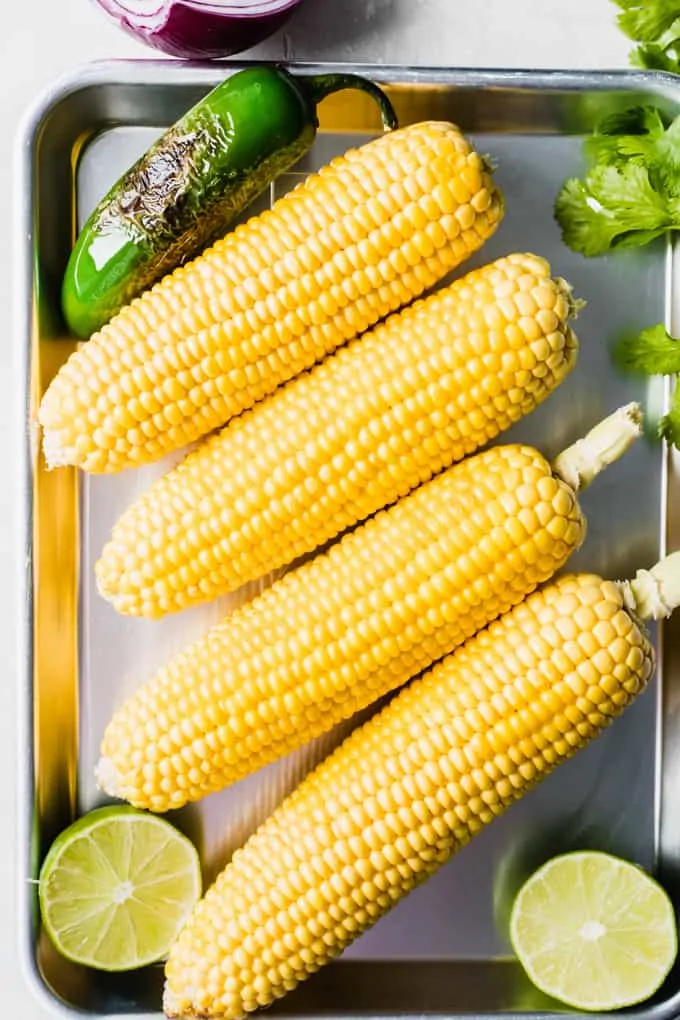 Small baking tray filled with cooked corn on the cob, roasted jalapeno, lime wedges and fresh cilantro.