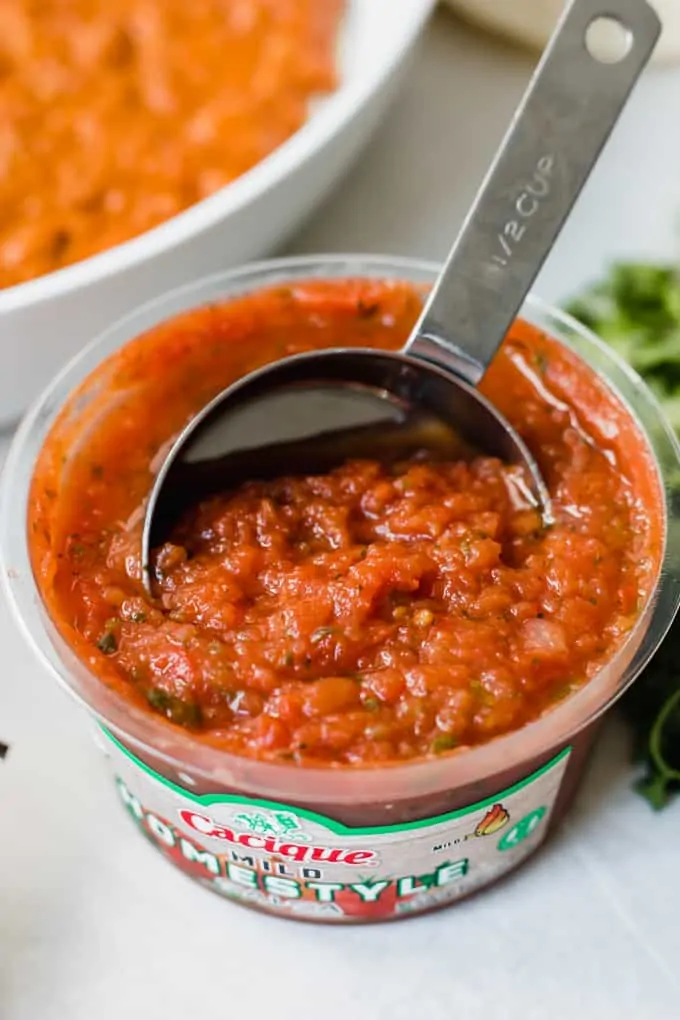 A measuring cup scooping up Cacique Homestyle Salsa.