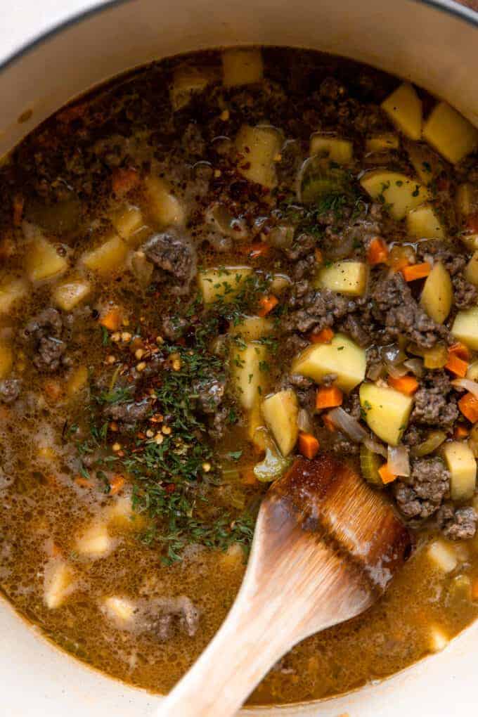 Dutch oven filled with bits of cooked ground beef, diced potatoes in a colorful broth and topped with herbs and seasonings to make the BEST cheeseburger soup.