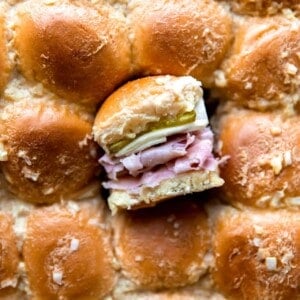 Overhead shot of the tops of sliders with one slider cut out and turned on it's side to show the layers of buns, sliced ham, Swiss cheese and pickles on the inside.