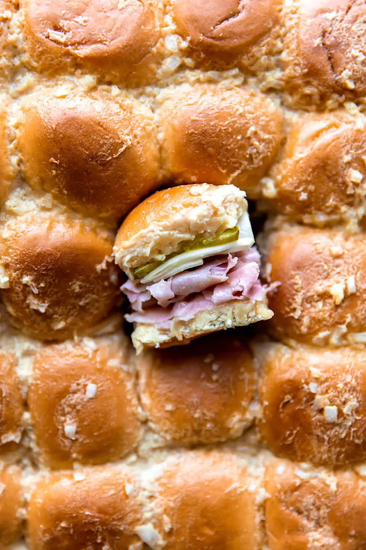 Overhead shot of the tops of sliders with one slider cut out and turned on it's side to show the layers of buns, sliced ham, Swiss cheese and pickles on the inside.