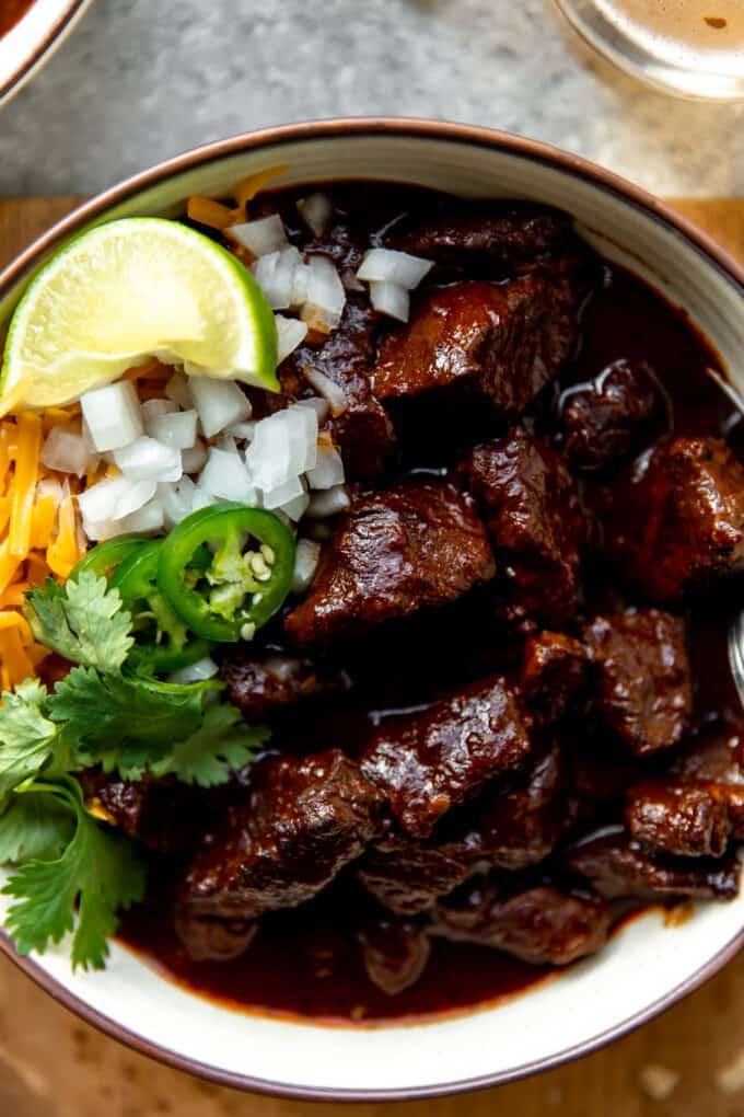 Bowl replaced with Authentic Texas Chili, showing thick chunks of meat covered in a thick red sauce. Coberto com cebola em bico, queijo picado, coentros, jalapeño fatiado e uma cunha de Lima.