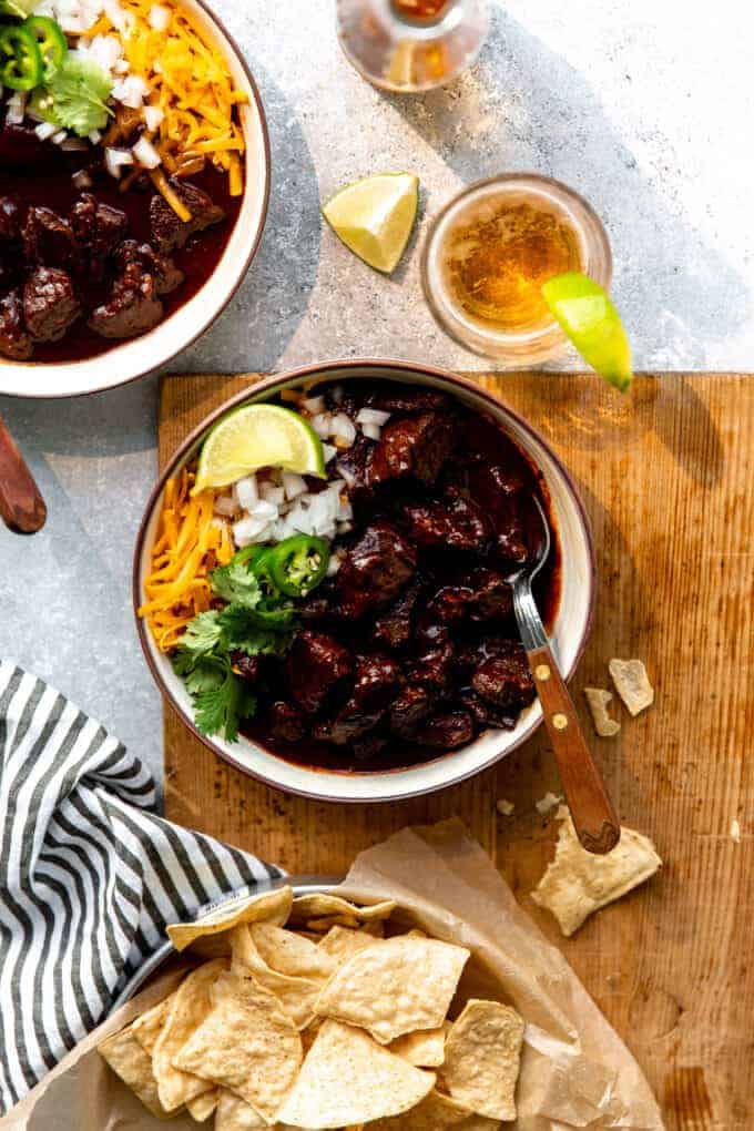 Overhead showing a table setting with homemade True Texas Chili, bowls of chili served with tortilla chips on the side, glasses of beer with lime wedges and a bottle.