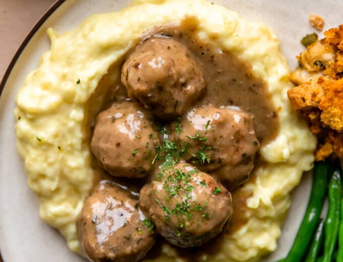 Turkey meatballs and gravy served over mashed potatoes on a plate with green beans and stuffing on the side.